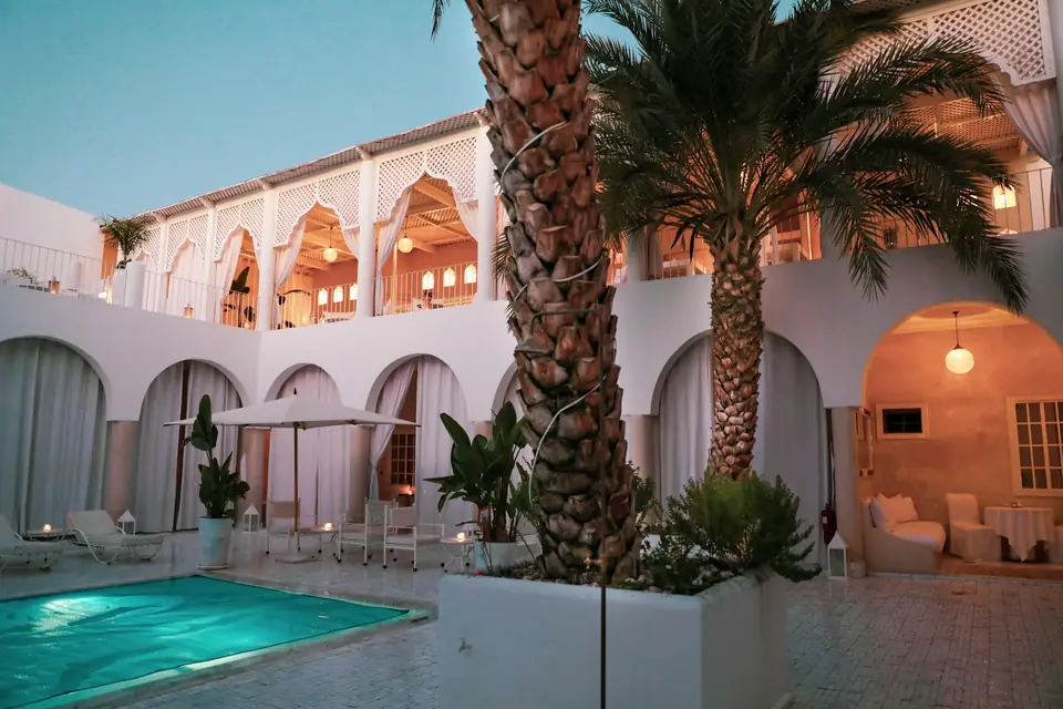 Cour de riad blanche à arcades, piscine et palmiers à la tombée du jour